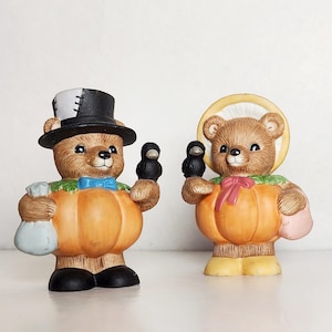 May include: Two ceramic bear figurines dressed as pumpkins for Halloween. One bear is wearing a black top hat and holding a small black bird. The other bear is wearing a yellow headband and holding a small black bird.