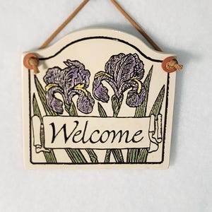 May include: A ceramic welcome sign featuring two purple iris flowers and green leaves. The sign has a cream-colored background with black outlines and the word "Welcome" in a banner. A brown string is attached for hanging.