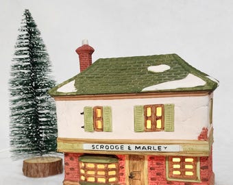 May include: A ceramic Christmas village building with a green roof and red brick accents. The building has the text "Scrooge & Marley" on a sign above the door. A small green Christmas tree is to the left of the building.
