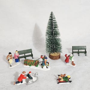 May include: A miniature Christmas village scene with figurines, including people, a Christmas tree, benches, and a sled. The figurines are painted with bright colors and depict winter activities.