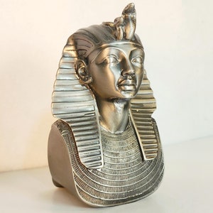 May include: A silver-colored bust of an Egyptian pharaoh, featuring a detailed headdress with a cobra. The bust has a serene facial expression and is set against a white background. The pharaoh's collar and headpiece are intricately designed.