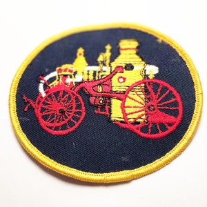 May include: A yellow and red embroidered patch depicting a vintage fire engine on a dark blue background. The patch has a yellow border.
