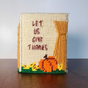 May include: A rectangular plastic canvas needlepoint decoration featuring the text LET US GIVE THANKS in brown yarn. The design includes a bright orange pumpkin, yellow accents, and a textured gold yarn curtain detail on the right side.