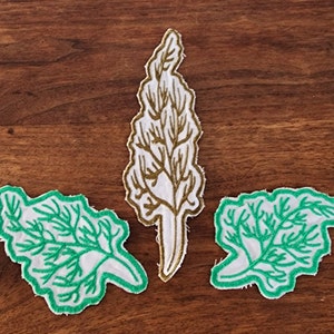 May include: Three embroidered fabric patches featuring green leafy designs on a white background. The center patch is a tall, thin leaf with gold embroidery. The other two patches are smaller and have a more rounded shape.