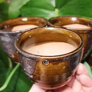 May include: Three brown ceramic mugs with a dark brown glaze and a light brown rim. The mugs are filled with a light brown liquid.