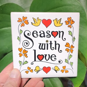 May include: White ceramic tile with a black and red design. The tile features a stylized floral border with red hearts and yellow birds. The text reads "Season with love."