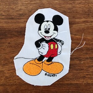 Peut inclure: Un patch en tissu blanc avec une image brodée de Mickey Mouse. Mickey porte son short rouge et ses chaussures jaunes emblématiques. Le patch est signé "©DISNEY".