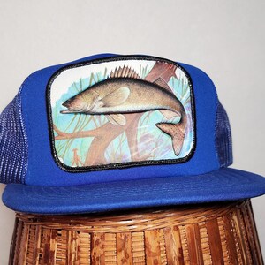May include: Blue and black mesh trucker hat with a patch of a fish on a branch. The patch is white with brown and green details.