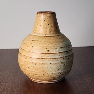 May include: A small, handcrafted ceramic vase with a rounded body and a narrow neck. The vase is a light beige color with brown speckles and horizontal brown lines. The vase is likely used for displaying flowers or as a decorative item.