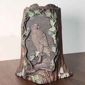 May include: A decorative, cylindrical container shaped like a tree stump. The front features a relief carving of a hawk perched on a branch, framed by vines with green leaves. The container is painted in shades of brown, gray, and green.
