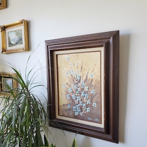 May include: A framed painting of a bouquet of blue flowers with white centers. The flowers are painted in a textured style, giving the painting a 3D effect. The frame is dark brown wood with a simple design.