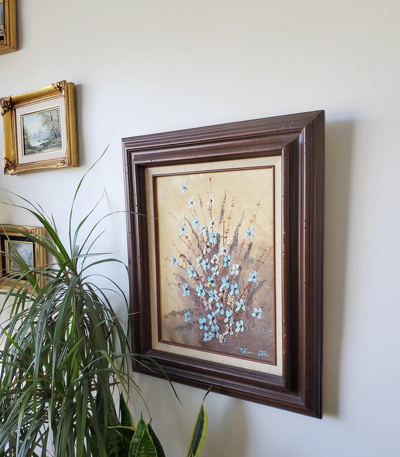 May include: A framed painting of a bouquet of blue flowers with white centers. The flowers are painted in a textured style, giving the painting a 3D effect. The frame is dark brown wood with a simple design.