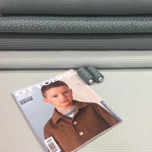 May include: A stack of four different shades of green waffle weave fabric with white polka dot fabric in the middle. A sewing magazine titled "Ottobre Design" with a young boy on the cover is on top of the fabric. Two spools of green thread are next to the magazine.