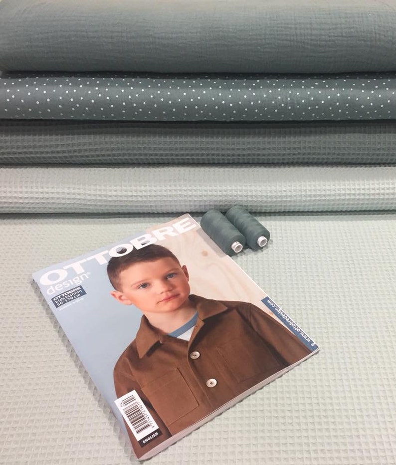 May include: A stack of four different shades of green waffle weave fabric with white polka dot fabric in the middle. A sewing magazine titled "Ottobre Design" with a young boy on the cover is on top of the fabric. Two spools of green thread are next to the magazine.