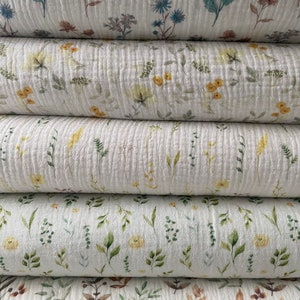 May include: A close-up of several stacks of white, textured fabric with a floral pattern. The fabric features watercolour-style illustrations of various flowers and foliage in shades of blue, yellow, green, and brown. The fabric appears to be a lightweight material.