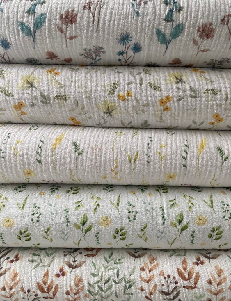 May include: A close-up of several stacks of white, textured fabric with a floral pattern. The fabric features watercolour-style illustrations of various flowers and foliage in shades of blue, yellow, green, and brown. The fabric appears to be a lightweight material.