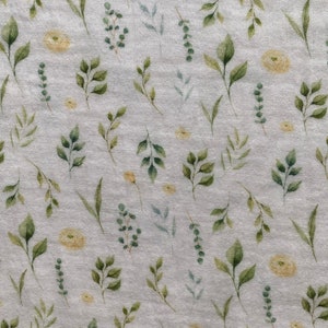 May include: A light grey fabric featuring a watercolour pattern of green leaves and small yellow flowers. The leaves vary in shape and size, creating a natural, botanical design. The fabric has a slightly textured appearance.