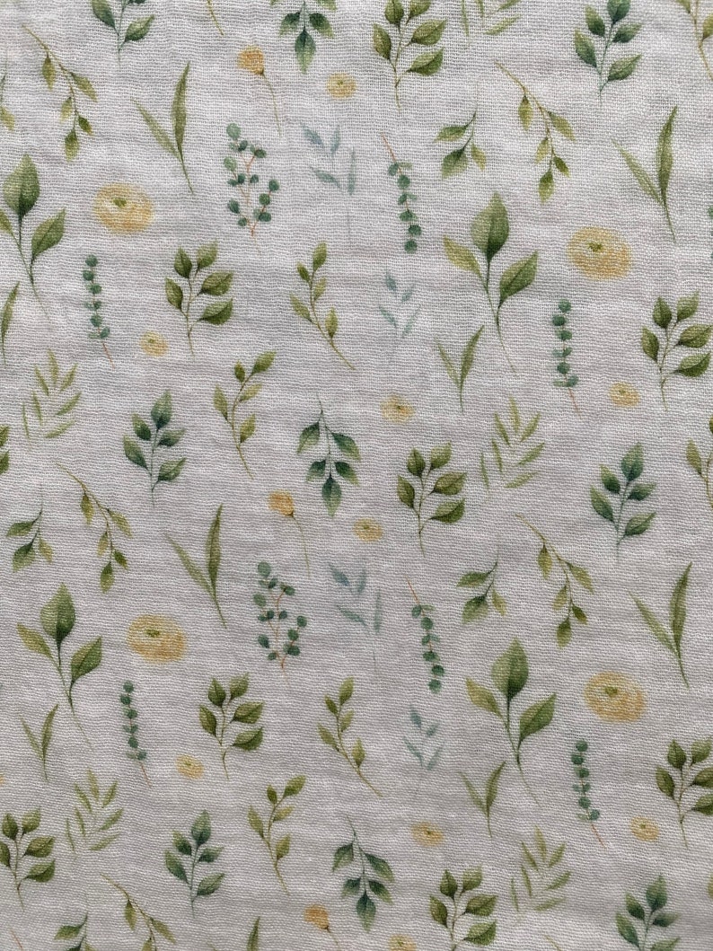 May include: A light grey fabric featuring a watercolour pattern of green leaves and small yellow flowers. The leaves vary in shape and size, creating a natural, botanical design. The fabric has a slightly textured appearance.