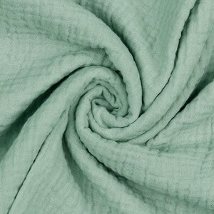 May include: A close-up of a soft, sage green fabric with a crinkled texture. The fabric is folded and twisted, creating a swirling pattern.