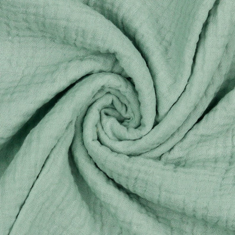 May include: A close-up of a soft, sage green fabric with a crinkled texture. The fabric is folded and twisted, creating a swirling pattern.