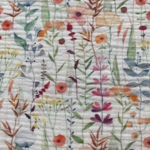 May include: Close-up of a white fabric with a watercolour floral pattern. The design features a variety of flowers in shades of orange, pink, red, and purple, along with green leaves and brown stems. The fabric has a textured, crinkled appearance.