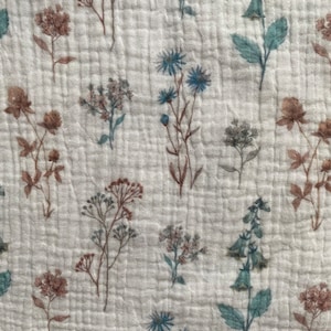 May include: Close-up of a white fabric with a floral pattern. The design features brown, blue, and green flowers, creating a delicate, botanical pattern. The fabric has a subtle, quilted appearance.