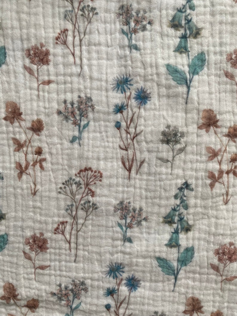 May include: Close-up of a white fabric with a floral pattern. The design features brown, blue, and green flowers, creating a delicate, botanical pattern. The fabric has a subtle, quilted appearance.