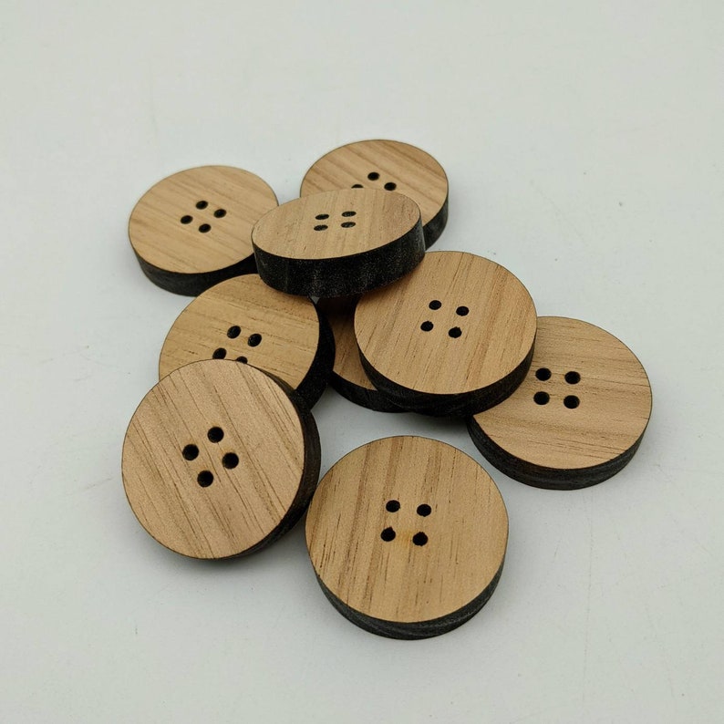 May include: A group of eight wooden buttons with a dark brown edge and five holes in the centre. The buttons are arranged in a pile on a white surface.