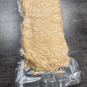 May include: A clear plastic bag filled with a large amount of light brown, shredded wood. The bag is sealed and has a label with the text "RP" on it.