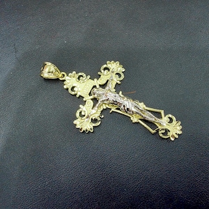 Op de afbeelding: Een goudkleurige kruisvormige hanger met een gedetailleerd ontwerp. Het kruis heeft een figuur van Jezus in zilverkleurig metaal. De hanger heeft een sierlijk, filigraanachtig ontwerp en een lus voor een ketting.