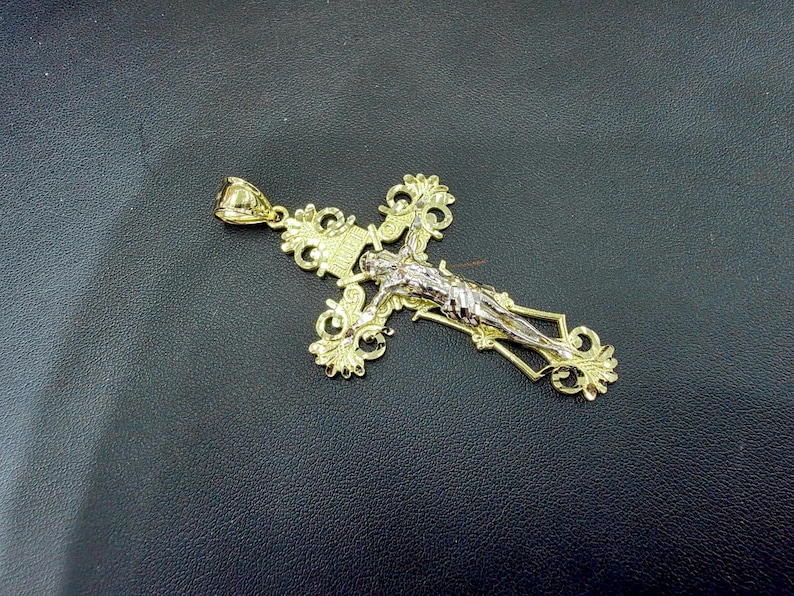 Op de afbeelding: Een goudkleurige kruisvormige hanger met een gedetailleerd ontwerp. Het kruis heeft een figuur van Jezus in zilverkleurig metaal. De hanger heeft een sierlijk, filigraanachtig ontwerp en een lus voor een ketting.