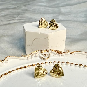 May include: Gold-colored heart-shaped stud earrings with a textured surface. Two pairs of earrings are displayed, one pair on a white block and the other on a white decorative plate with gold trim. The earrings are a warm, metallic tone.