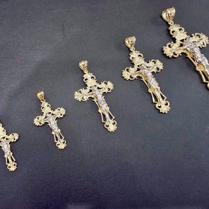 Op de afbeelding: Vijf goudkleurige kruisvormige hangers met ingewikkelde ontwerpen en een zilverkleurige Jezusfiguur. De kruisen vari&euml;ren in grootte, met sierlijke details op de armen en een lus voor een ketting. De hangers worden op een donkere ondergrond getoond.