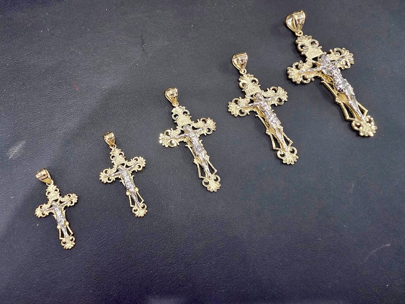 Op de afbeelding: Vijf goudkleurige kruisvormige hangers met ingewikkelde ontwerpen en een zilverkleurige Jezusfiguur. De kruisen vari&euml;ren in grootte, met sierlijke details op de armen en een lus voor een ketting. De hangers worden op een donkere ondergrond getoond.