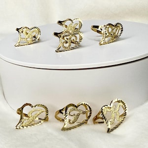 Puede incluir: Anillos dorados en forma de corazón con iniciales. Los anillos se exhiben sobre una superficie blanca. Cada anillo presenta un diseño de filigrana con una letra en el centro. Los anillos son de color dorado brillante.