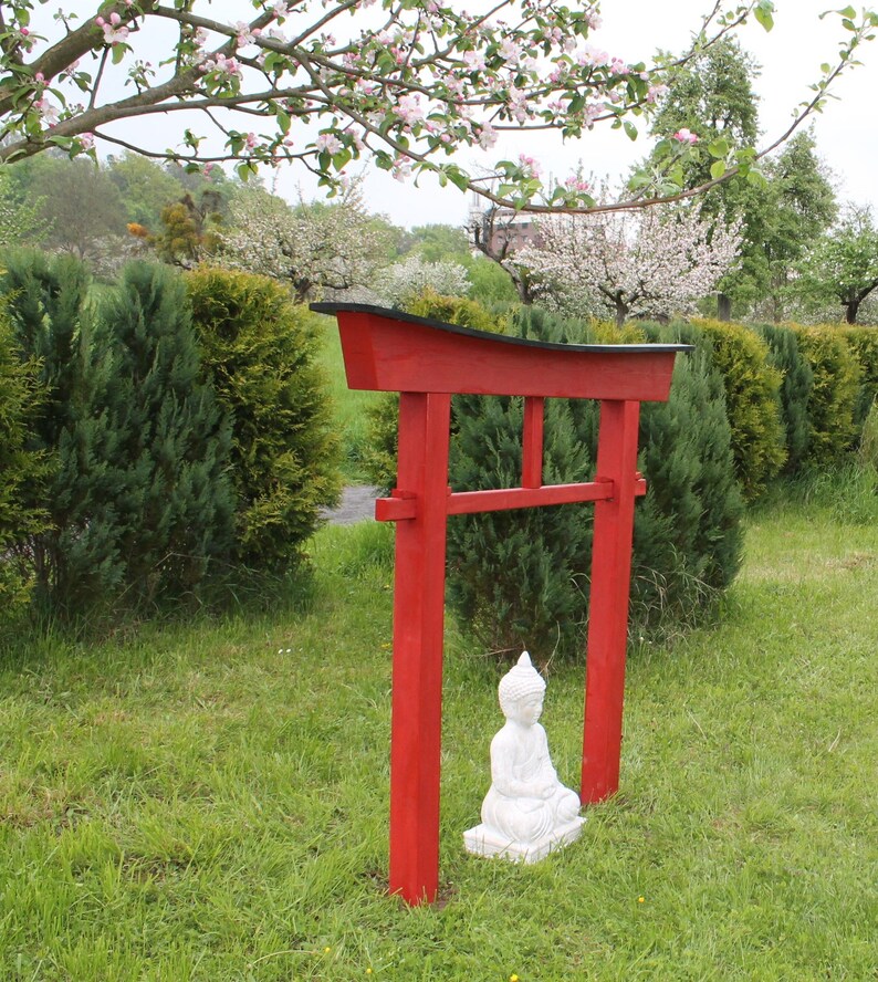 Pu&ograve; includere: Un torii in legno rosso con la parte superiore nera, in piedi in un'area erbosa con una statua bianca di un Buddha in meditazione seduto di fronte ad essa.