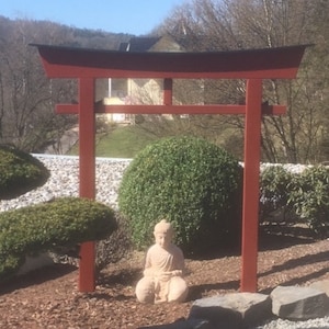 Pu&ograve; includere: Un torii in legno rosso con la parte superiore nera, in piedi in un giardino con una piccola statua di Buddha seduta di fronte ad esso.