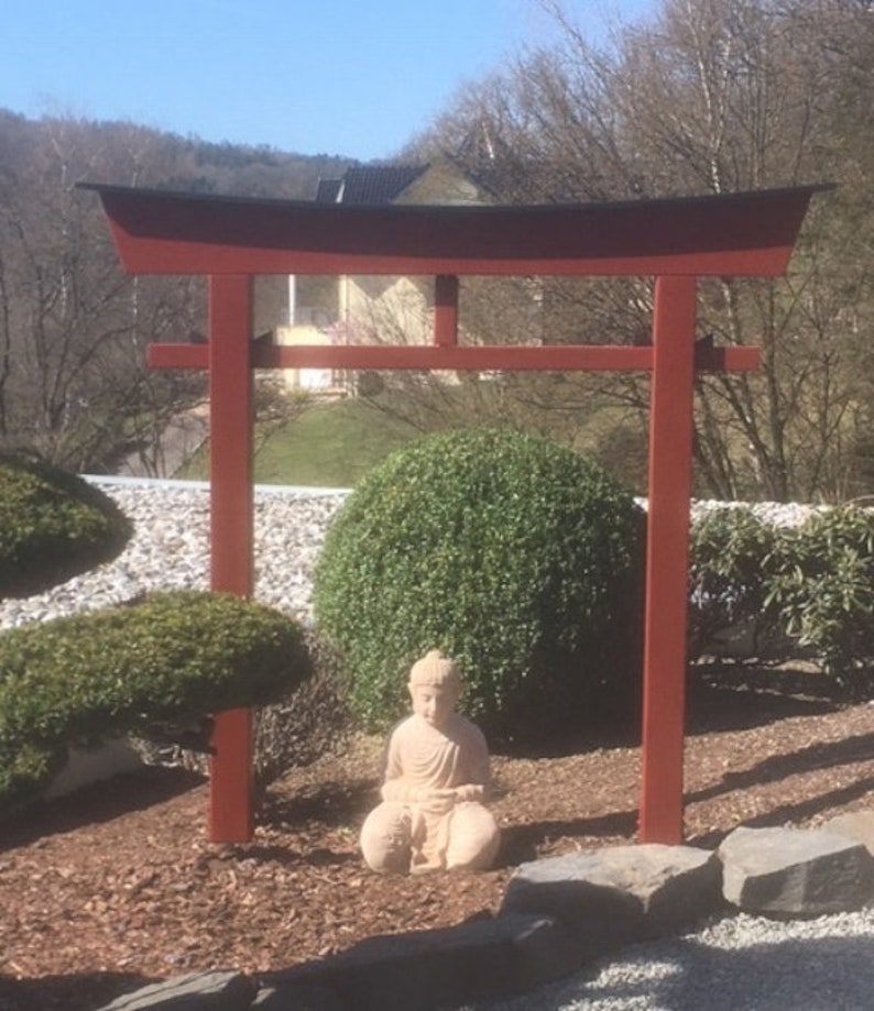 Pu&ograve; includere: Un torii in legno rosso con la parte superiore nera, in piedi in un giardino con una piccola statua di Buddha seduta di fronte ad esso.