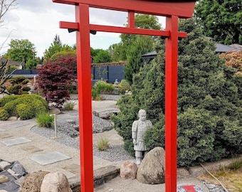 Può includere: Un alto portale torii rosso brillante caratterizzato da due montanti verticali e una traversa superiore curva con un bordo superiore nero. La struttura si erge in un giardino, incorniciando una vista di arbusti verdi, ghiaia e pietre.