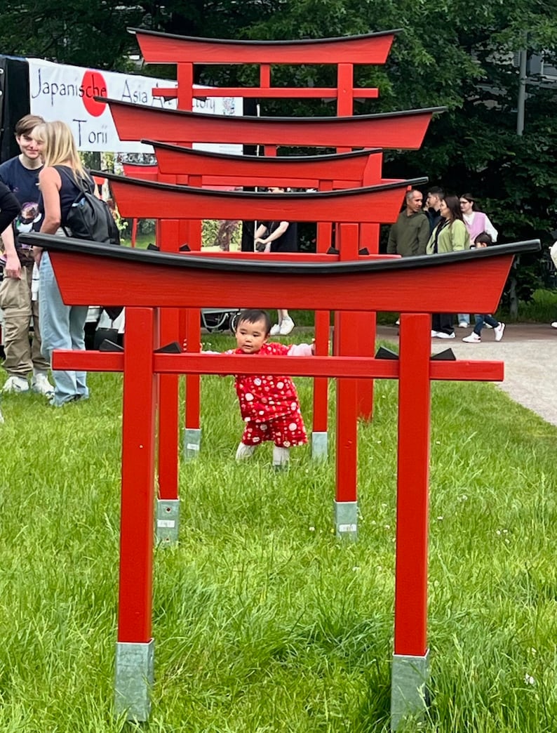 Pu&ograve; includere: Una bambina con un vestito a pois rosso e bianco cammina attraverso una serie di torii rossi. I torii sono installati in un'area erbosa con vegetazione verde sullo sfondo.