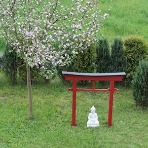 Pu&ograve; includere: Un torii in legno rosso con una statua bianca di una figura in meditazione seduta di fronte ad esso. Il torii &egrave; situato in un'area erbosa con un albero fiorito alle sue spalle.