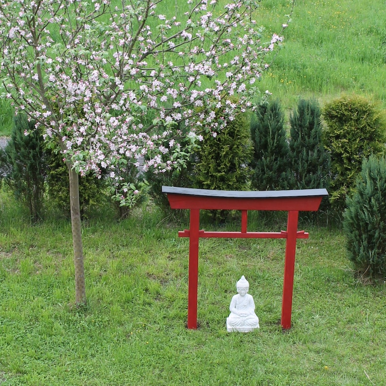 Pu&ograve; includere: Un torii in legno rosso con una statua bianca di una figura in meditazione seduta di fronte ad esso. Il torii &egrave; situato in un'area erbosa con un albero fiorito alle sue spalle.