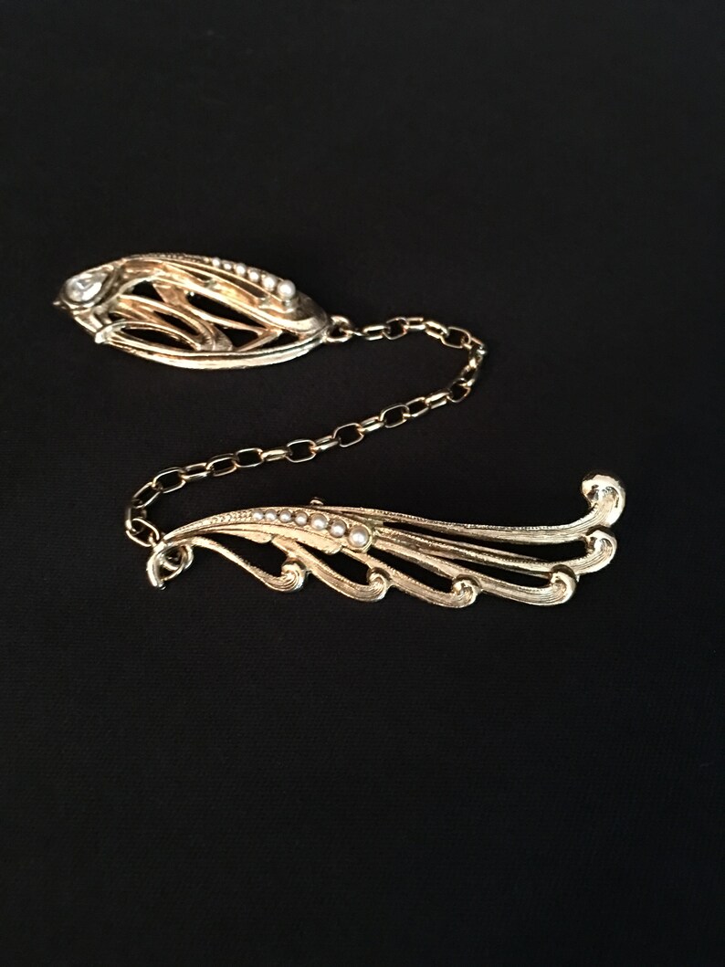 One side is oval seed pearls and rhinestone dramatic sweater pins Vintage gold 4\u201d chain between them the other is a swoosh