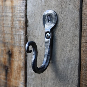 May include: A hand-forged, black metal hook fixed to a wooden surface. The hook has a curved design with a decorative top, and is secured with a screw. The top of the hook has the letter "A" stamped on it.