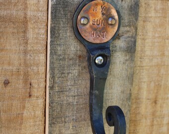 May include: A handcrafted, black metal hook with a copper plate that reads "MY SUN HAT" attached to a wooden surface. The hook has a decorative, curled end. The wood has a natural, weathered appearance.