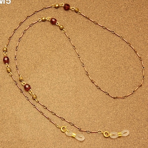May include: A gold-coloured eyeglass chain with a delicate design. The chain features small, round, red beads interspersed with smaller gold beads. The chain is designed to hold eyeglasses and has clear rubber loops at each end.