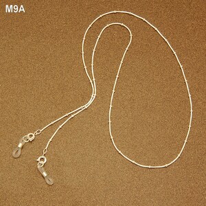 May include: A silver glasses chain with small, evenly spaced beads. The chain has loops at each end to attach to glasses frames. The chain is displayed on a brown surface. The text "M9A" is in the upper left corner.