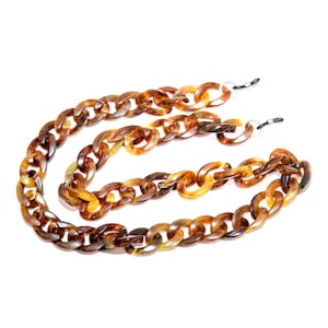 May include: A tortoiseshell-patterned chain-link eyeglass holder. The chain is made of large, rounded links in shades of brown and tan. The ends have small loops for attaching to glasses. The chain is shown against a white background.