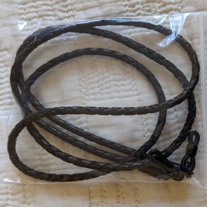 May include: A dark brown, plaited leather glasses cord, presented in a clear plastic bag. The cord has a loop at one end and a clasp at the other for attaching to spectacles. The plaited design provides a textured, stylish appearance.