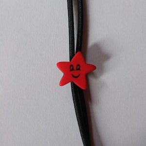 Può includere: Un elastico per capelli a forma di stella rossa con una faccia sorridente. La stella è centrata su un elastico nero. Lo sfondo è una superficie bianca.